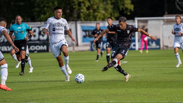 A photo that shows a football player passing the ball during a game. In the background the referee is sprinting to keep up with play.